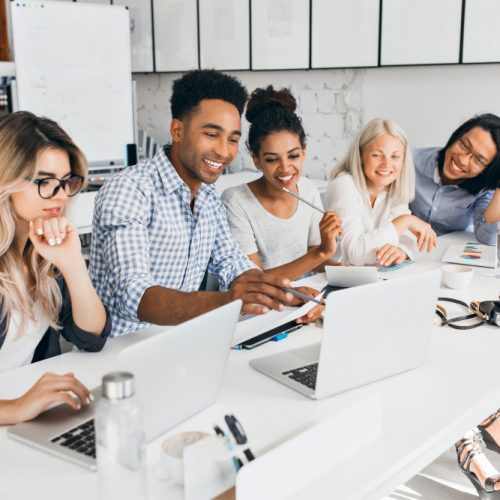 smiling-african-student-pointing-with-pencil-laptop-screen-concentrated-blonde-woman-glasses-propping-chin-with-hand-while-working-with-computer-office